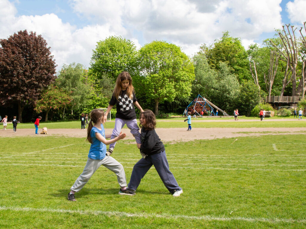 Three students build a human pyramid on a school field, with other children playing behind them