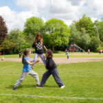 Three students build a human pyramid on a school field, with other children playing behind them