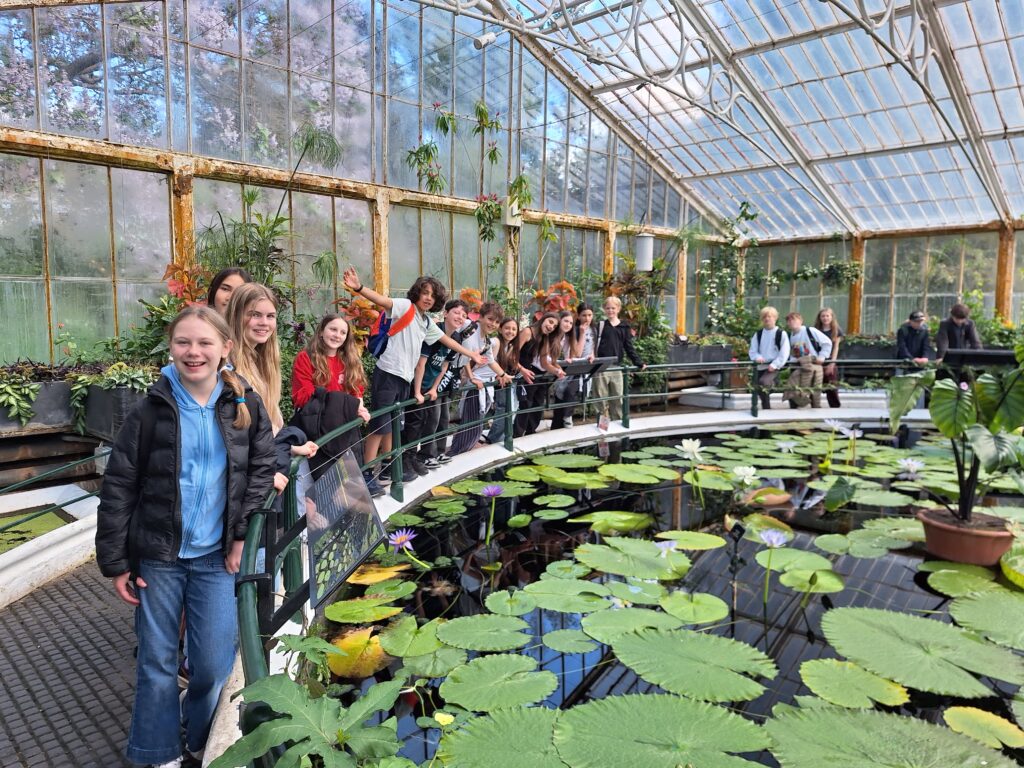 Students pose for a photo around a pond with lily pads on the surface inside a glasshouse