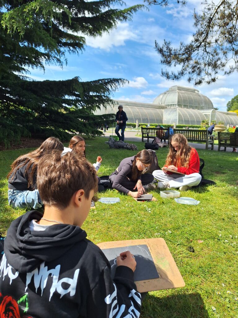 Students sit on grass and sketch, with a large glass house behind them