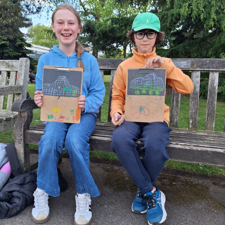 A female and male student sit on a bench, holding up sketches they have done of a large structure