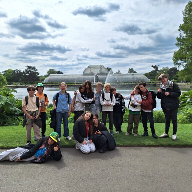 Students pose for a photo on a grassy verge. Behind them is a large lake with a large glasshouse on the far bank.