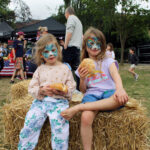 Two girls with their faces painted sit on a haystack eating burgers in buns on the field at The King Alfred School, North London