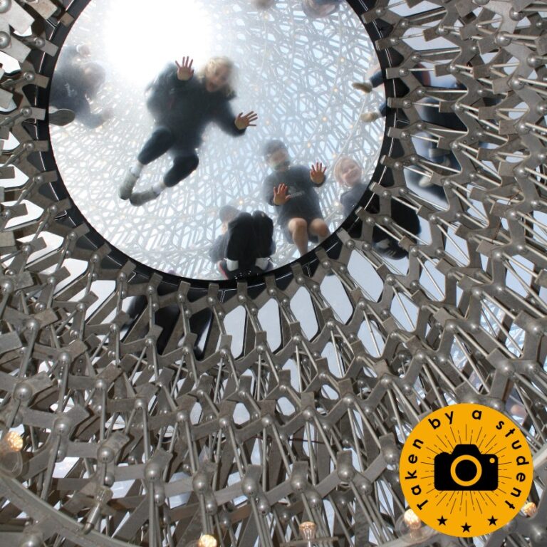Students appear blurry through a glass circle set in an architectural metal structure, pictured from below
