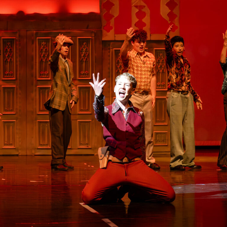 A male student signs passionately whilst on his knees during a performance of Sister Act Jr. Fellow performers are in the background.