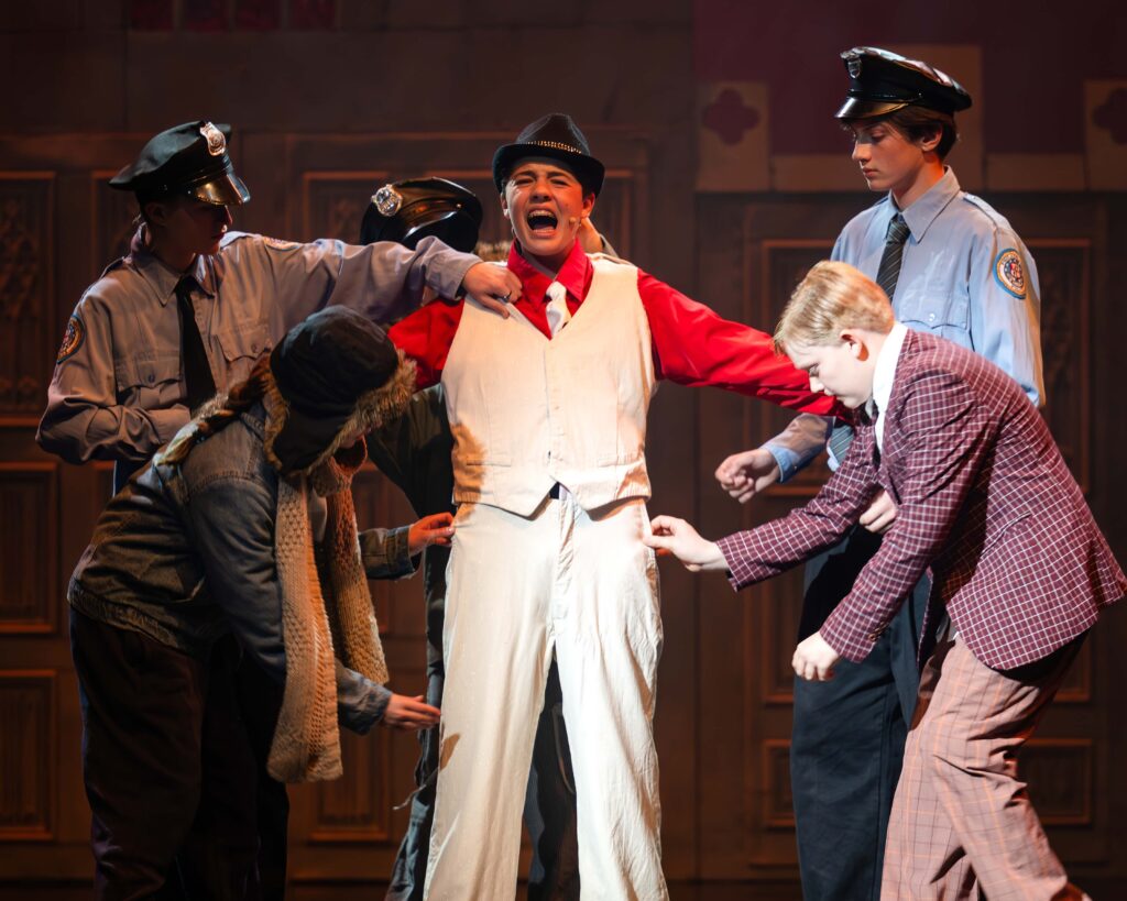 A male student dressed as a mobster is surrounding by fellow student actors on stage during a performance of Sister Act Jr
