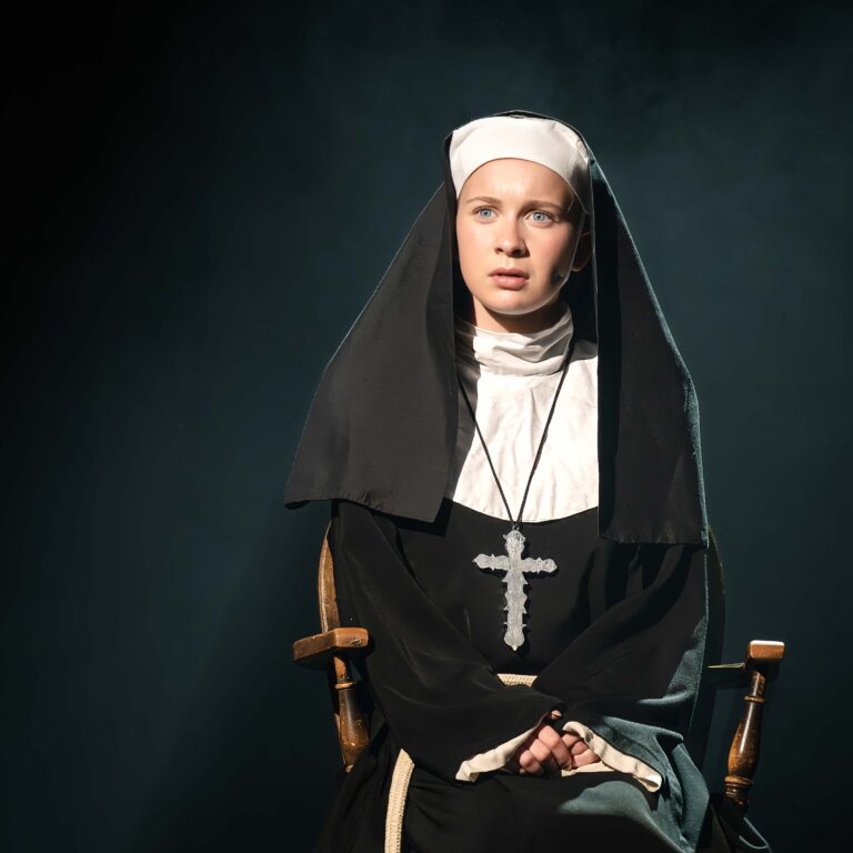 A lone female student dressed as a nun looks troubled sitting in a chair on a dark stage during a performance of Sister Act Jr