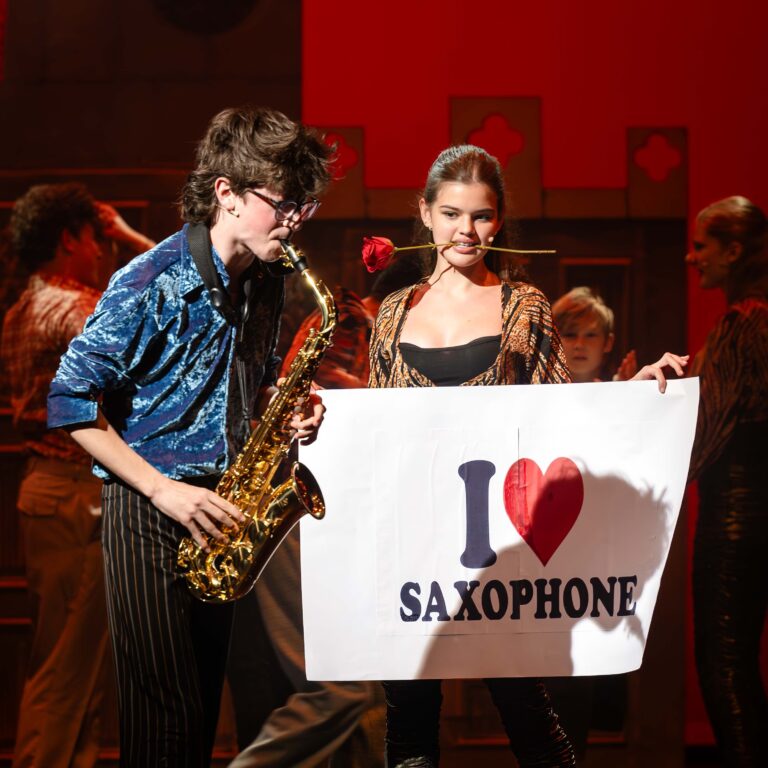 A male student plays saxophone enthusiastically whilst a female student holds a sign reading 'I heart saxophone' during a performance of Sister Act Jr