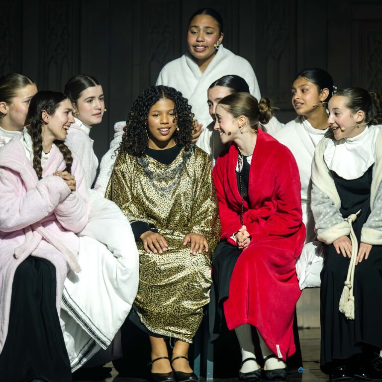 Female students in dressing gowns huddle around the lead actress on stage in The King Alfred School's productions of Sister Act Jr