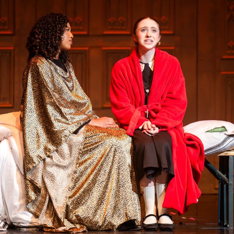 Two female students sit on stage, one wearing leopard print and the other in a red dressing gown, during a performance of Sister Act Jr