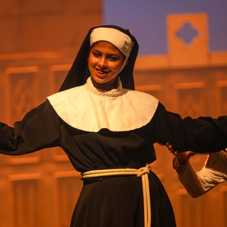 A female student dressed as a nun smiles and sings on stage with her arms outstretched
