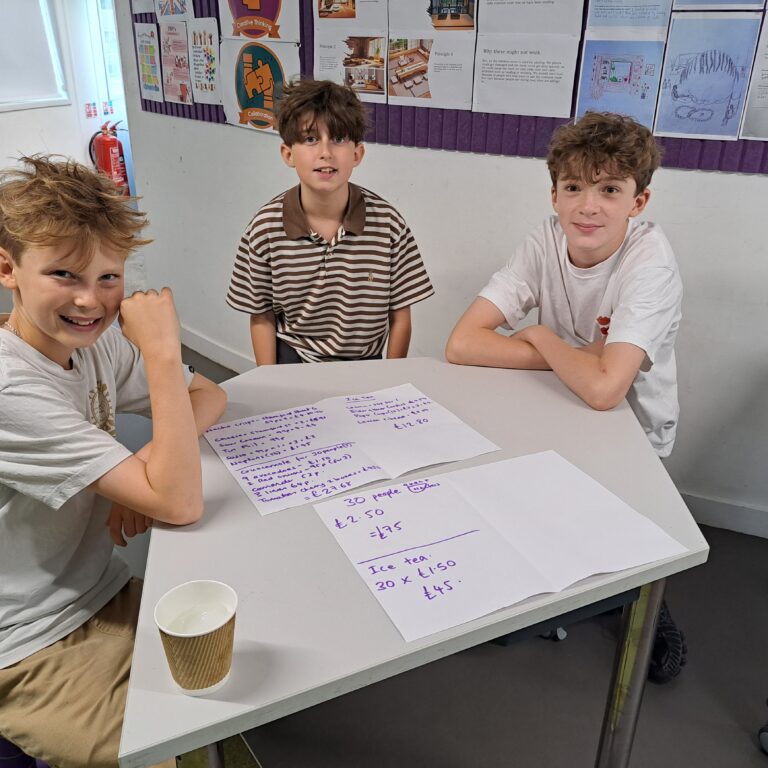Three male students sit at a table with the cost calculations for an iced tea stall on the desk in front of them