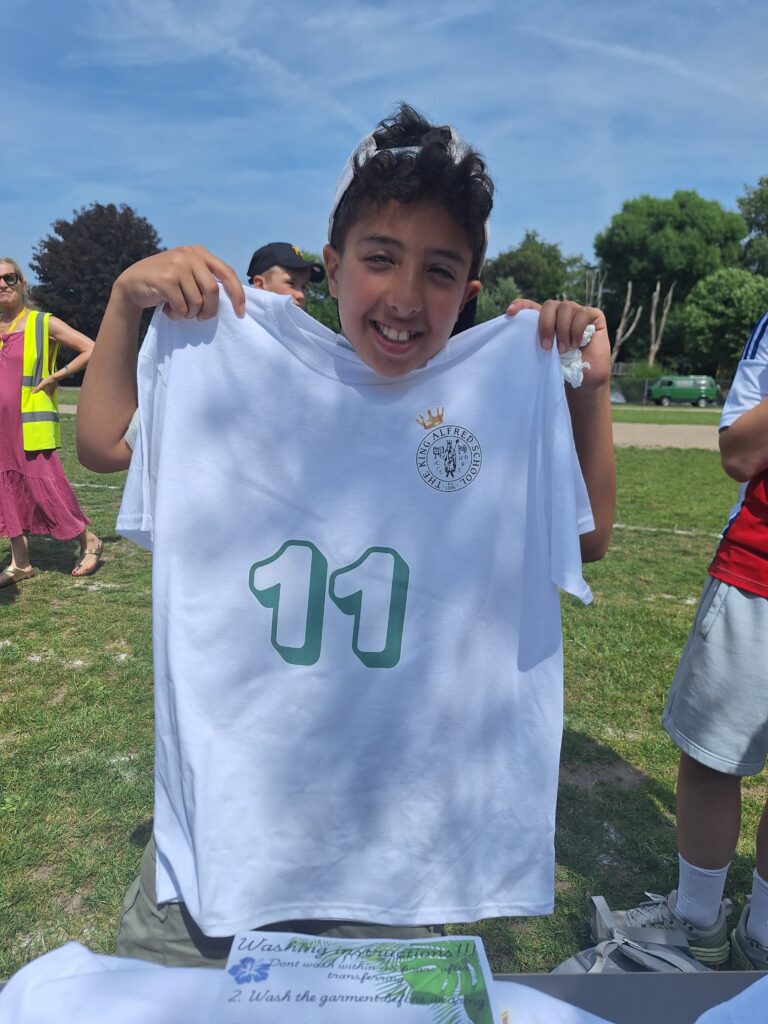 A male Year 7 student holds up a t-shirt with The King Alfred School logo and the number 11 printed on it, with a school field in the background