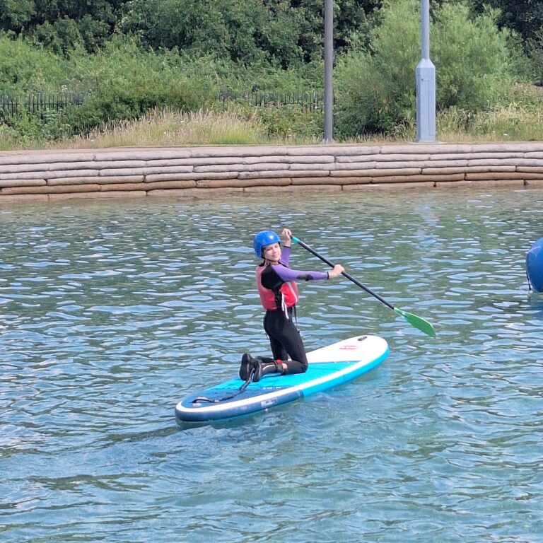 A student looks over their shoulder whilst holding an oar on a paddleboard in water