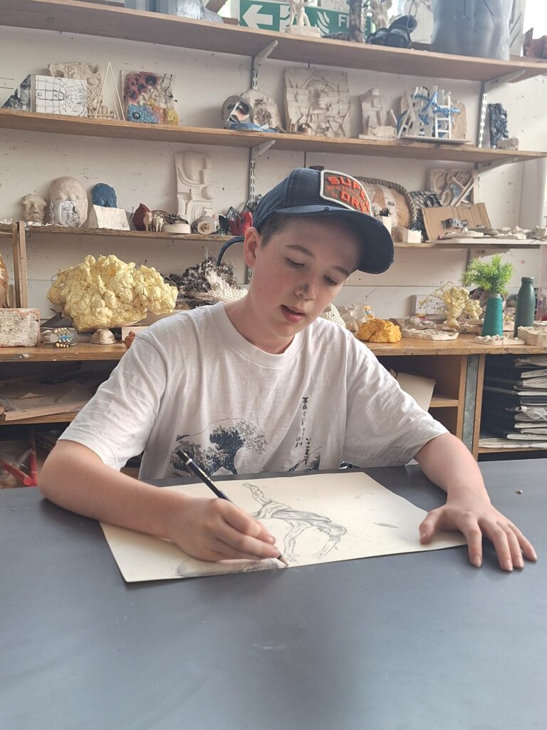 A male student sits in an art room sketching with a pencil and paper