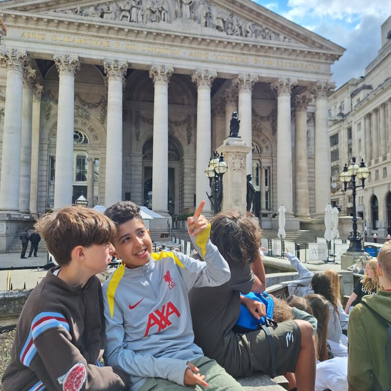 Students from The King Alfred School sit in front of the Royal Exchange building in London. One points at something whilst another looks on.