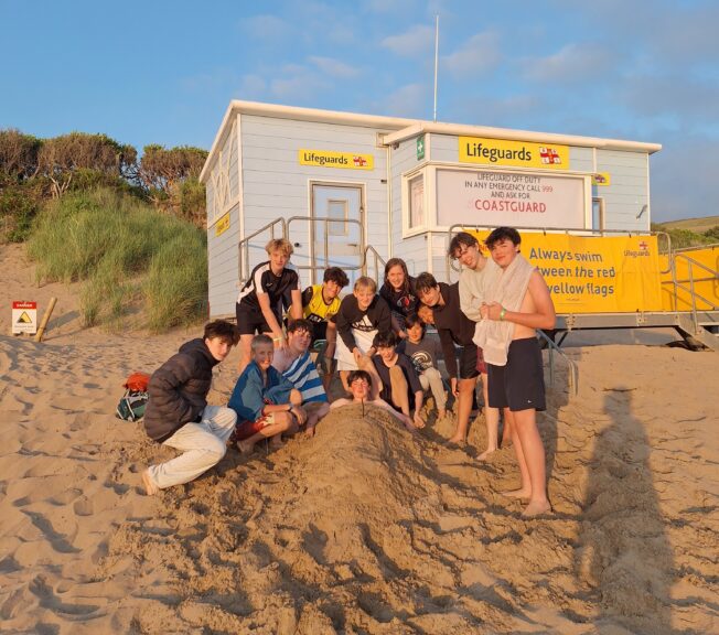 A group of students from The King Alfred School sit on a beach, with one student buried in the sand up to his neck. Sand dunces and a lifeguard station can be seen behind them.