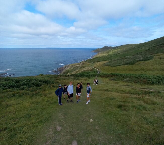A group of students walk a coastal path across a green clifftop, with the sea and a vast sky to the left and above.