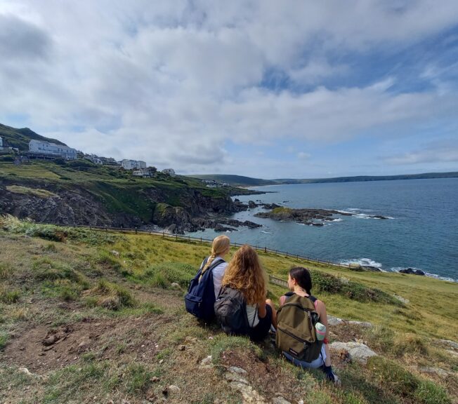 Three female students sit with their backs to the camera on a picturesque clifftop, with the sea beyond them.