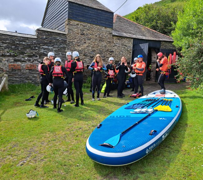 A group of 12 students wear wetsuits and stand next to a paddleboard on the floor. An adult instructor is giving a tutorial.