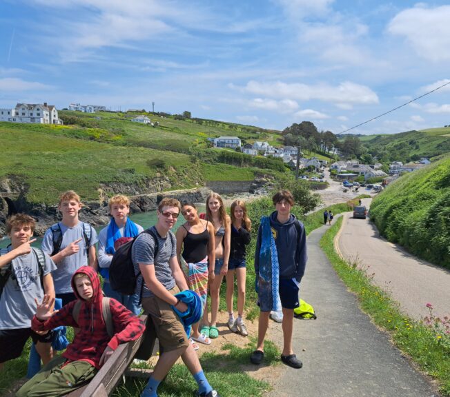 A group of nine students stand on a country lane, with green fields either side and a grand building in the distance.