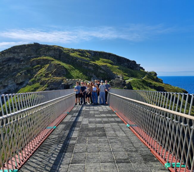 A group of students stand on a suspension bridge, with a clifftop, the sea and a vast blue sky behind them