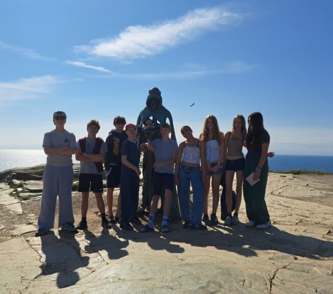 A group of nine students stand around a metal statue on a stone-platform by the sea
