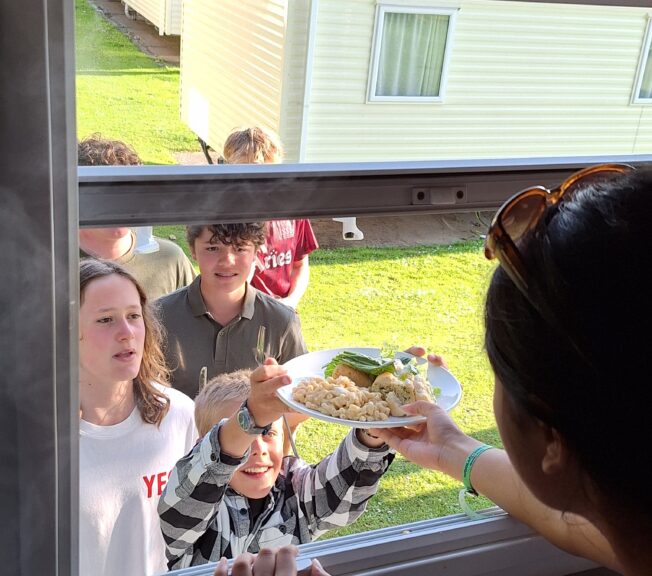 A girl in shadow hands a plate of food to a boy through the window of a static caravan. A queue of fellow students is stood behind him.