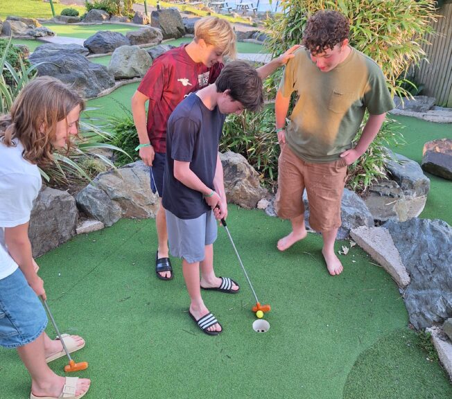 A group of four male students stand on a miniature golf course. One is putting a ball with a golf club, whilst the others watch on.