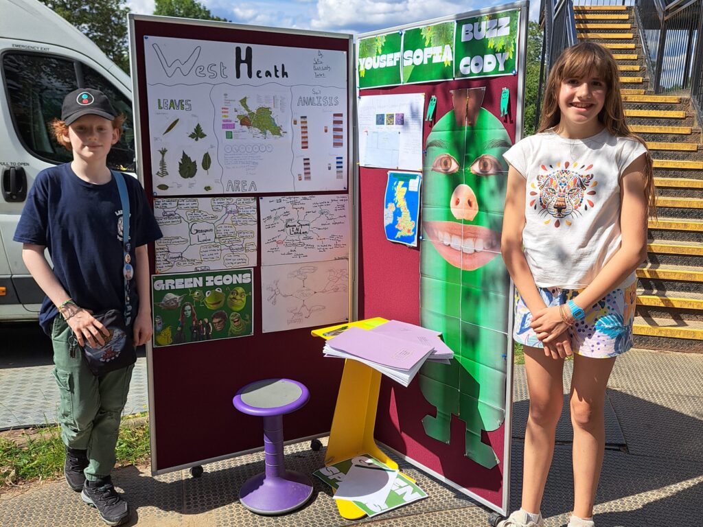 A smiling male and female student stand beside an outdoor display of their work charting the biodiversity of Hampstead Heath