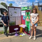 A smiling male and female student stand beside an outdoor display of their work charting the biodiversity of Hampstead Heath