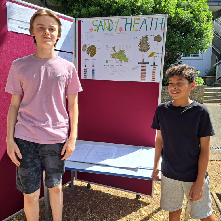 Two smiling male students stand beside an outdoor display of their work charting the biodiversity of Hampstead Heath