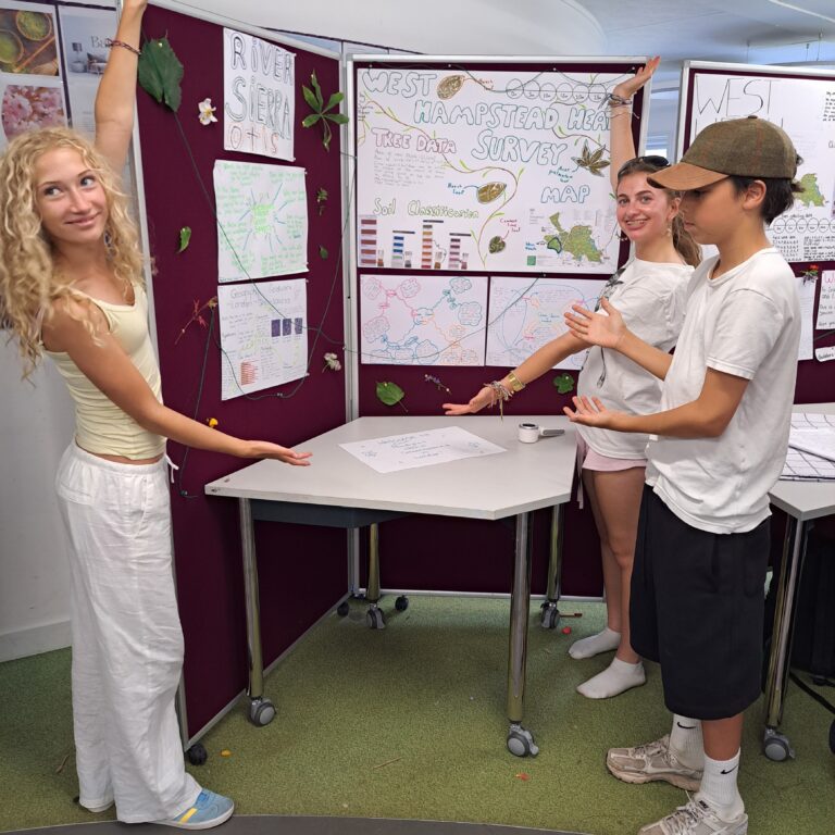 Three smiling students - two girls and a boy - stand with their arms outstretched, drawing attention to a display of their work charting the biodiversity of Hampstead Heath