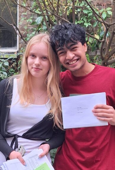 A male and female student smile and hold their GCSE results outside