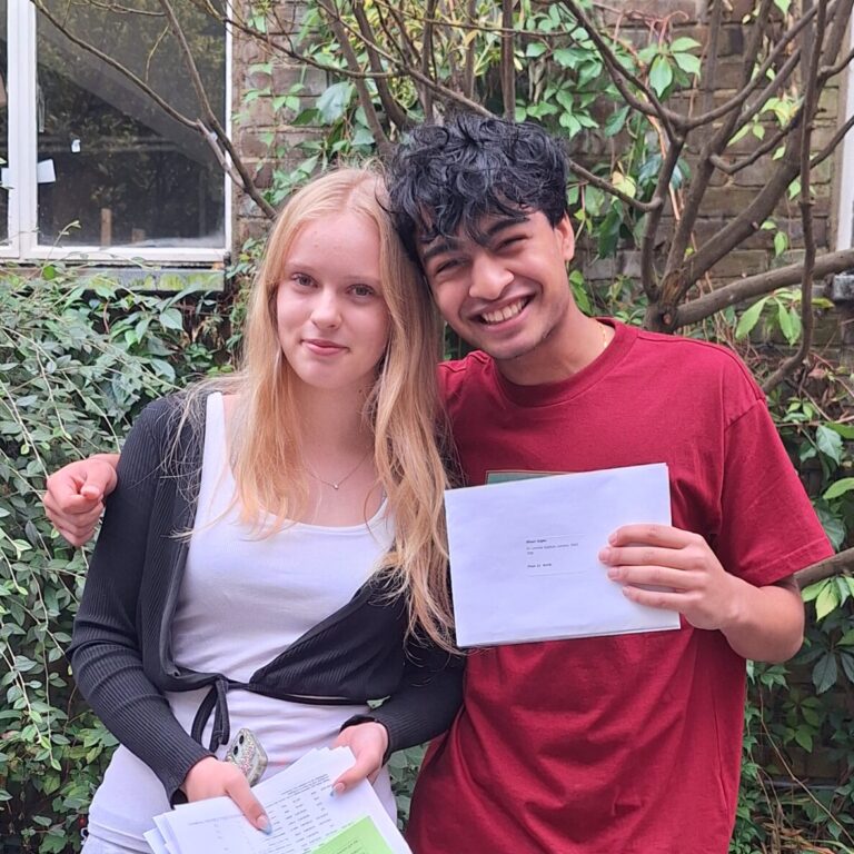 A male and female student smile and hold their GCSE results outside