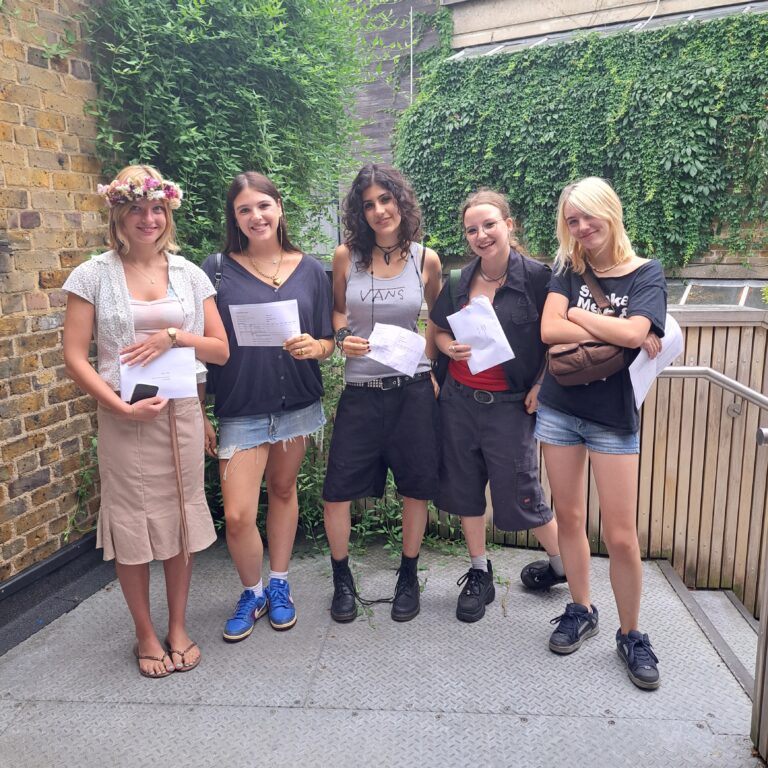 A group of five female students stand in a line outside. They are smiling and holding pieces of paper bearing their A level exam results.