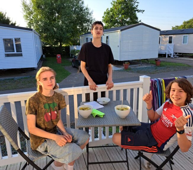 Three male students on the terrace of a static home at sundown. Two are sitting around a table and one stands.