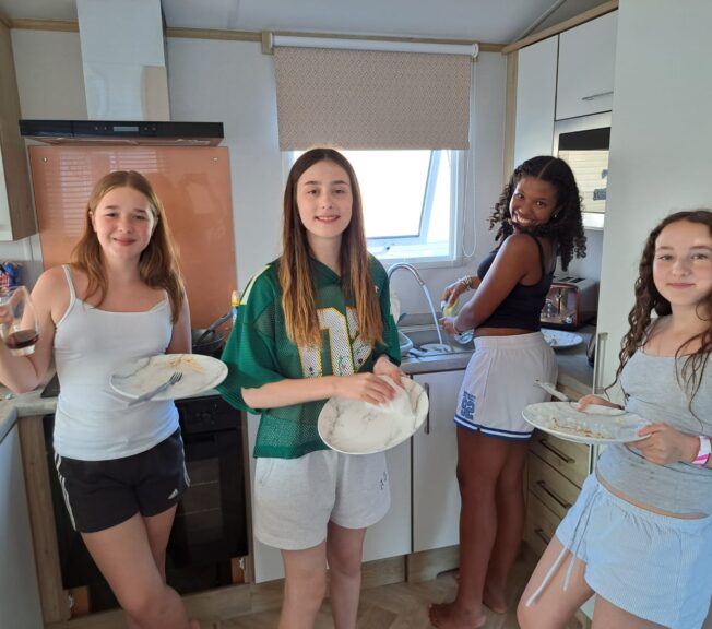 Four female students stand in a line in the kitchen of a static caravan. Two hold dirty dishes, one is washing up, and the fourth is using a tea towel to dry a plate.