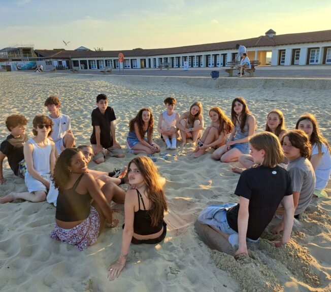 A group of 15 students sit in a circle on the beach at sundown. They are all turned to look at the camera. A long, low row of buildings can be seen behind them.