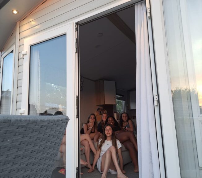 An open glass door on the side of a holiday home reveals a group of female students sat inside, smiling for the camera