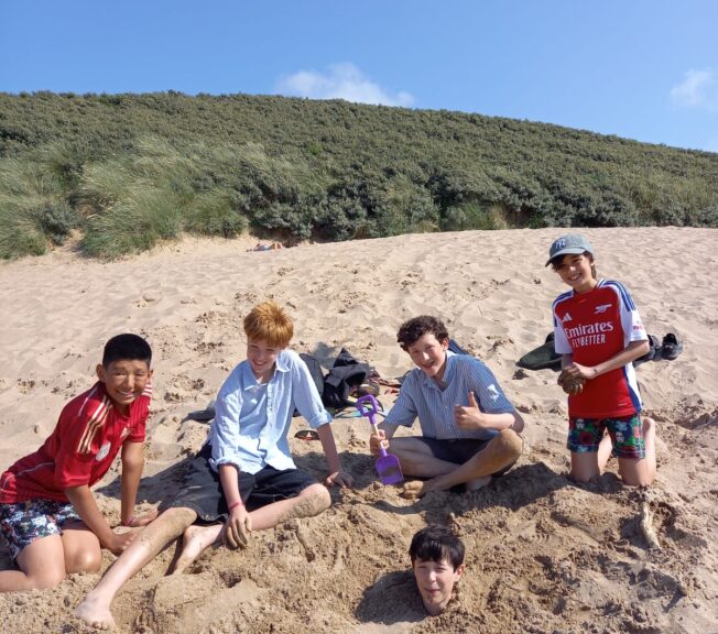 Four male students sit smiling on a sandy beach. a fifth boy can be seen buried in the sand, with only his head visible.
