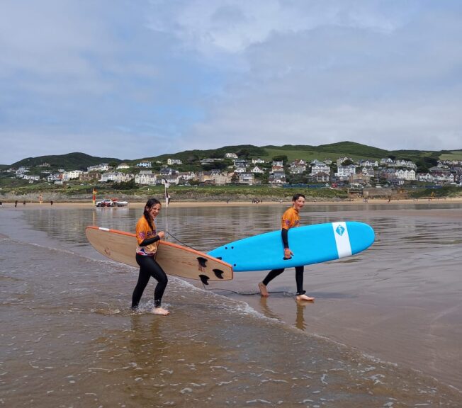 Two students wearing wetsuits carry surf boards across wet sand at a beach, with a small town and clifftops behind them.