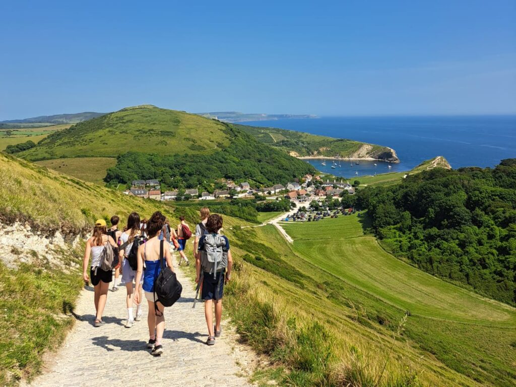 Students are pictured from behind walking down a coastal path, with greenery around them, the sea to the right and a big blue sky