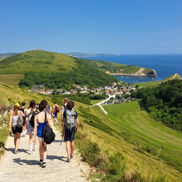Students are pictured from behind walking down a coastal path, with greenery around them, the sea to the right and a big blue sky
