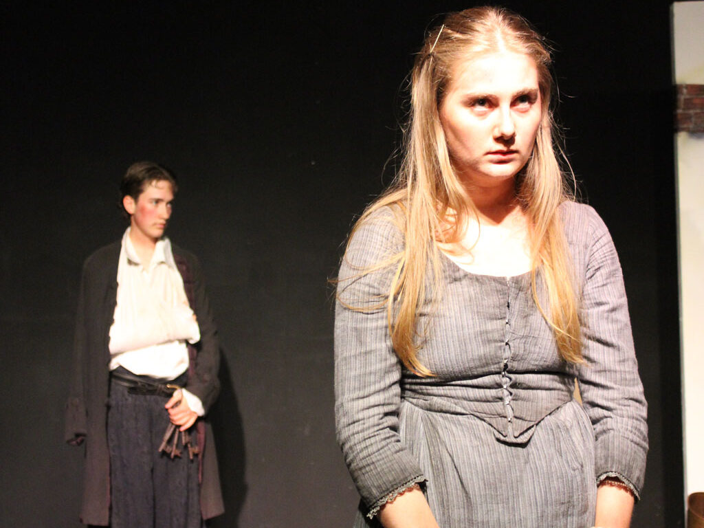 UAL Performing Art students from The King Alfred School wear period costume whilst performing a dramatic scene from The Welkin on stage. The protagonist looks forlorn in the foreground whilst another actor with their arm in a sling watches on behind.