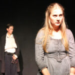 UAL Performing Art students from The King Alfred School wear period costume whilst performing a dramatic scene from The Welkin on stage. The protagonist looks forlorn in the foreground whilst another actor with their arm in a sling watches on behind.