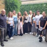 A group of nine people standing outside in a semi-casual setting, smiling for the camera. On the far left is Robert Lobatto, Head of The King Alfred School North London. The rest are students dressed casually in jeans, skirts, or trousers. They are standing against a background of a brick wall on the left, a wooden panel on the right, and a green ivy-covered wall behind them.