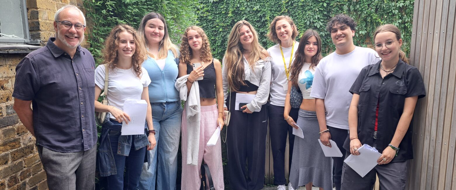 A group of nine people standing outside in a semi-casual setting, smiling for the camera. On the far left is Robert Lobatto, Head of The King Alfred School North London. The rest are students dressed casually in jeans, skirts, or trousers. They are standing against a background of a brick wall on the left, a wooden panel on the right, and a green ivy-covered wall behind them.