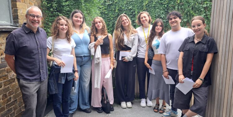 A group of nine people standing outside in a semi-casual setting, smiling for the camera. On the far left is Robert Lobatto, Head of The King Alfred School North London. The rest are students dressed casually in jeans, skirts, or trousers. They are standing against a background of a brick wall on the left, a wooden panel on the right, and a green ivy-covered wall behind them.
