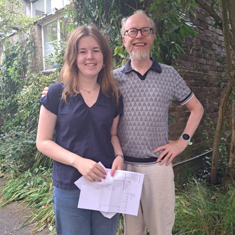 A female student holds her exam results on a piece of paper, standing next to a male teacher beaming with pride. Behind them is lots of greenery.